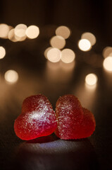 Two red sugar marmalade candies in shape of heart on black background, lit from side, behind round yellow blurred lights