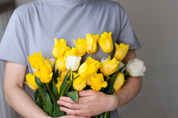 A woman in a gray dress holds a large bouquet of yellow tulips. Beautiful background with spring yellow flowers, give a bouquet, a gift. Photo with bright colors, Mother's Day, March 8, Valentine's