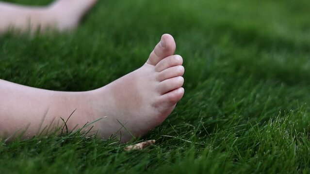 Close-up of the baby little bare foots on green soft grass. Infant sitting and playing on lawn at the park outdoors in summer warm day. First steps concept. Happy children day, happy childhood