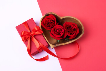 Three red rosebuds in a wooden bowl in the shape of a heart with red gift box on a pink and red background. Happy Valentine's Day, Mother's Day and birthday greeting card.