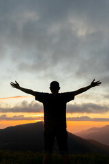 silhouette of young man standing on top of mountain peak with his hands raised above his head as symbol of worship and invocation of God's blessings with belief and  power of faith in God. - obrazy, fototapety, plakaty
