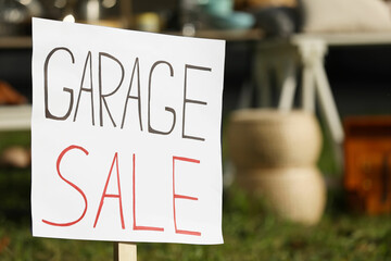 Sign Garage sale written on cardboard near tables with different stuff outdoors, closeup. Space for text
