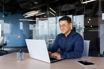 Successful mature asian working inside office using laptop, man typing on keyboard and smiling,...