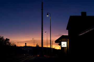 Evening photo of an empty train station. Colorful sky and railway lights. The clock reads 5:40 p.m. - obrazy, fototapety, plakaty