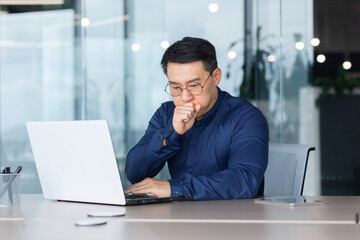 Asian businessman sick working at workplace inside office with laptop, man in shirt and glasses coughs and has a cold.