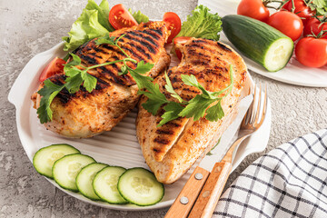 two large juicy healthy pieces of grilled chicken fillet on a white ceramic serving tray with fresh cucumber, parsley. diet lunch.