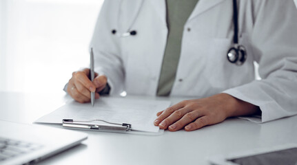 Unknown doctor woman sitting and writing notes at the desk in clinic or hospital office, close up.  Medicine concept.