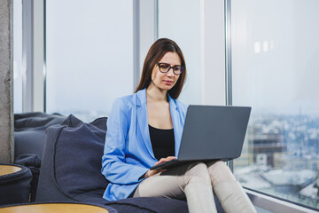 Business young woman in glasses with long dark hair in casual clothes smiling and looking at laptop while browsing documents online during weekend in workspace