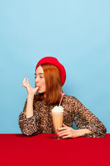 Sweet life. Young woman in red beret tasting delicious milkshake with whipped cream over blue studio background. Food pop art photography. Complementary colors. Concept of art, beauty, food. Ad