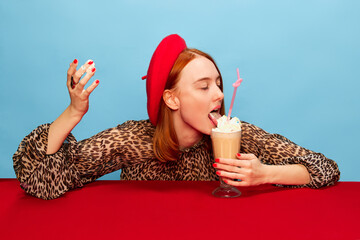 Tasting, licking whipped cream. Young woman in red beret drinking milkshake over blue studio background. Food pop art photography. Complementary colors. Concept of art, beauty, food. Copy space for ad