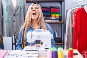 Blonde woman dressmaker designer using sew machine angry and mad screaming frustrated and furious, shouting with anger. rage and aggressive concept.