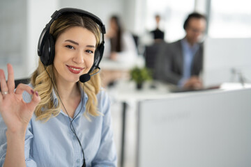 Portrait of beautiful blond haired woman with headset showing OK sing at call centre