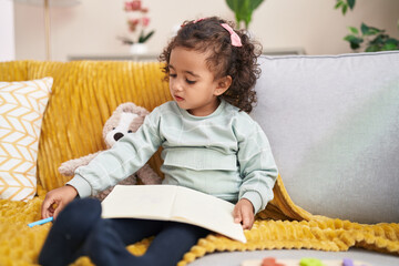 Adorable hispanic girl drawing on notebook sitting on sofa at home