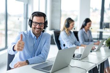 Handsome male online support agent with headset showing thumb up accompanied with his colleagues at...