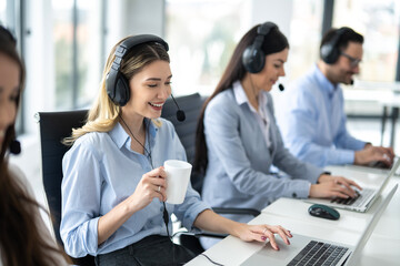 Fototapeta premium Smiling blond haired woman operator with headset drinking morning coffee and starting doing her online job at customer support center with her business colleagues.