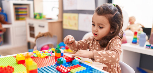 Adorable hispanic girl playing with construction blocks sitting on table at kindergarten