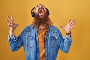 Caucasian man with long beard listening to music using headphones crazy and mad shouting and yelling with aggressive expression and arms raised. frustration concept.