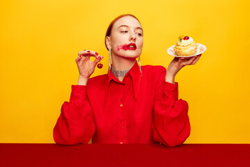 Beautiful young girl in red shirt tasting cake with cherry over yellow studio background. Food pop art photography. Complementary colors. Concept of art, beauty, food. Copy space for ad, text.