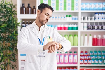 Handsome hispanic man working at pharmacy drugstore checking the time on wrist watch, relaxed and...