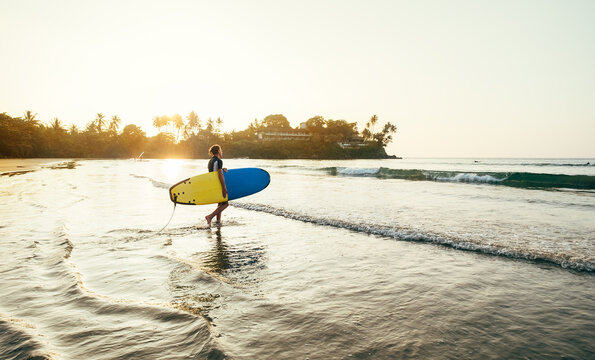 Teen boy with blue and yellow surfboard entering the waves for surfing with sunset rays on palm trees grove. Happy childhood and active sporty people vacation time concept.
