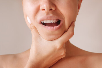 Cropped shot of a young woman suffering from jaw pain holding her chin isolated on a beige background. Inflammation of cervical lymph nodes, Diseases of ENT organs, facial, trigeminal nerve, toothache - obrazy, fototapety, plakaty