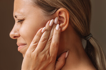 Young caucasian blonde woman holding her ears with her hands isolated on a dark brown background. Pain and tinnitus, ear otitis