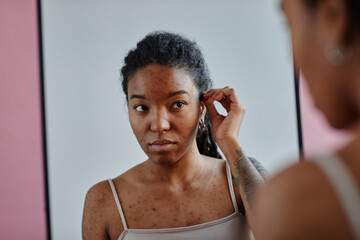 Young black woman with acne scars looking in mirror insecure in appearance