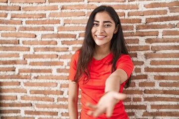 Young teenager girl standing over bricks wall smiling friendly offering handshake as greeting and welcoming. successful business.