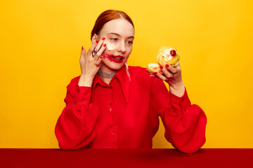 Surrealism. Beautiful young girl in red shirt with smudged red lipstick, eating cake over yellow studio background. Food pop art photography. Complementary colors. Concept of art, beauty, food.