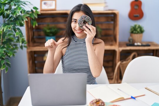 Young Teenager Girl Using Computer Laptop With Doughnut On Face Pointing Thumb Up To The Side Smiling Happy With Open Mouth