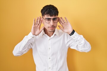 Young hispanic man standing over yellow background trying to hear both hands on ear gesture, curious for gossip. hearing problem, deaf