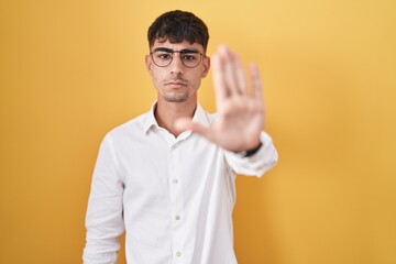 Young hispanic man standing over yellow background doing stop sing with palm of the hand. warning...