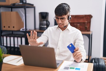 Young hispanic man working using computer laptop holding credit card doing stop gesture with hands...