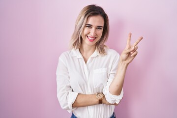 Young beautiful woman standing over pink background smiling with happy face winking at the camera doing victory sign with fingers. number two.