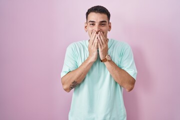 Handsome hispanic man standing over pink background laughing and embarrassed giggle covering mouth...