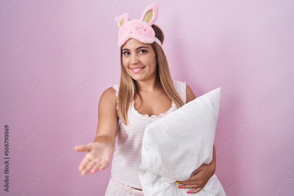 Sticker Young blonde woman wearing pyjama hugging pillow smiling cheerful offering palm hand giving assistance and acceptance.