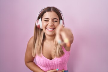 Young blonde woman listening to music using headphones laughing at you, pointing finger to the...