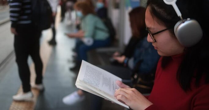 Young asian woman waiting at tram station while listening music playlist and reading book