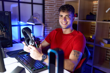 Young hispanic man streamer smiling confident holding headphones at gaming room