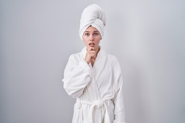Blonde caucasian woman wearing bathrobe looking fascinated with disbelief, surprise and amazed expression with hands on chin