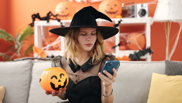 Young blonde woman holding halloween pumpkin basket using smartphone at home