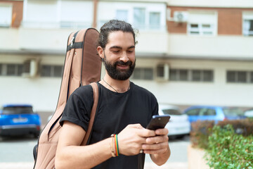 Young hispanic man musician using smartphone holding guitar case at park