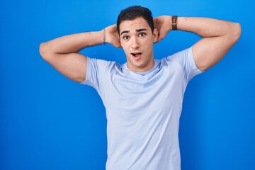 Young hispanic man standing over blue background crazy and scared with hands on head, afraid and surprised of shock with open mouth