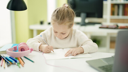 Adorable blonde girl student writing on notebook sitting on table at classroom