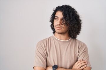 Hispanic man with curly hair standing over white background looking to the side with arms crossed convinced and confident