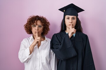 Hispanic mother and daughter wearing graduation cap and ceremony robe asking to be quiet with...