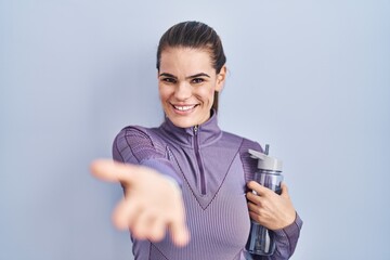 Beautiful woman wearing sportswear holding water bottle smiling cheerful offering palm hand giving assistance and acceptance.