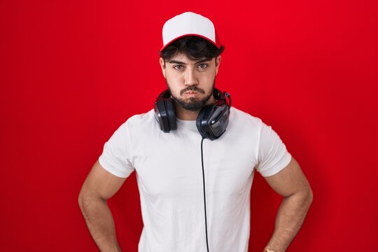 Hispanic man with beard wearing gamer hat and headphones puffing cheeks with funny face. mouth inflated with air, crazy expression.