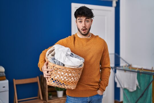 Hispanic man with beard holding laundry basket at laundry room scared and amazed with open mouth for surprise, disbelief face