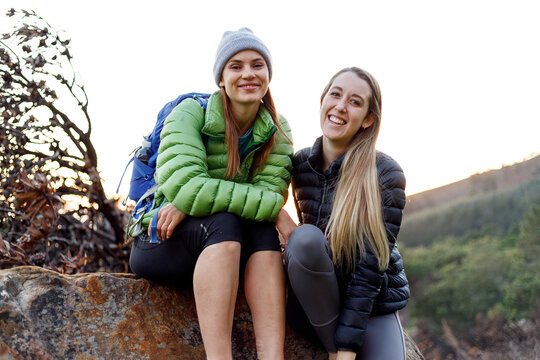 Smiling young woman in down jacket sitting with friend on rock with their pet dog